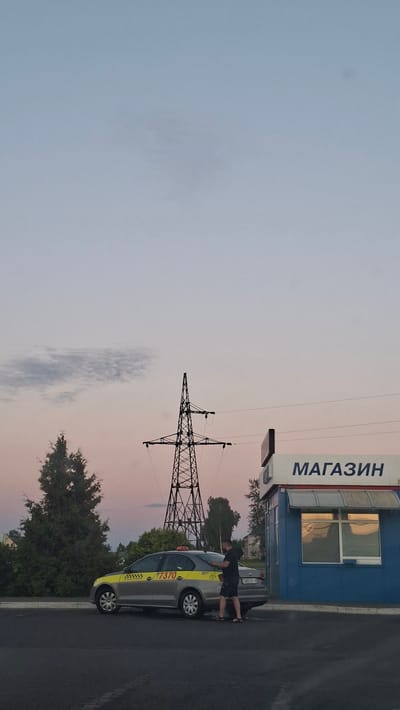 The image depicts an exterior scene at dusk, likely in the city of Smalyavichy, Belarus, given the presence of Cyrillic text on the building. In the foreground, a grey sedan taxi with yellow and grey livery, displaying the number "7370" and a license plate starting with "4991 A1-8", is parked. A man wearing dark shorts and a dark shirt is standing beside the rear passenger side of the taxi, appearing to interact with it. Behind the taxi, a blue building with a grey awning over a window is visible. The building has the word "МАГАЗИН" (which translates to "STORE" in Russian) written in large white letters across its facade. In the mid-ground, a tall electricity pylon stands against a twilight sky that transitions from pale blue at the top to a soft pinkish hue near the horizon. A few trees, including a coniferous tree, are situated to the left of the pylon. The overall environment suggests an urban or suburban setting during the early evening.