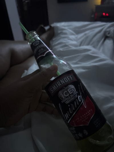A hand holds a partially consumed bottle of Smirnoff Ice Double Black with Guarana, a pre-mixed alcoholic beverage. A green straw protrudes from the bottle. The background features a white, rumpled fabric and what appears to be a nightstand. The location is not discernible as Port Harcourt, Nigeria without additional information.