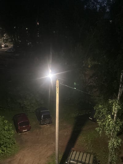 The image depicts an outdoor scene at night, illuminated by a bright streetlamp positioned on a horizontal pole. The scene appears to be a somewhat rural or suburban area, possibly a backyard or side street in Kirov, Russia, given the context.

Dominating the foreground is the intense glare of the streetlamp, creating a strong bloom of light that washes out much of the immediate surroundings. Below the light pole, a gravel or dirt path is visible, bordered by greenery and trees.

Two cars are parked on the path. The car closer to the foreground is a dark red sedan. Further up the path, a darker colored vehicle, possibly a hatchback or SUV, is parked. Both cars appear to be unoccupied. In the background, dense foliage and trees are present, with some appearing to be deciduous trees. The light from the lamp also casts shadows and highlights the textures of the leaves and branches. To the far right, a portion of a structure with a slatted roof or side is visible, possibly a shed or a utility structure.

The overall atmosphere is dark and somewhat mysterious due to the nighttime setting and the intense light source. No people are visible in the frame, and the scene suggests a quiet, uneventful moment. There is no discernible text in the image. The presence of the streetlamp suggests an area with public lighting, but the surrounding darkness and vegetation point towards a less urbanized setting.