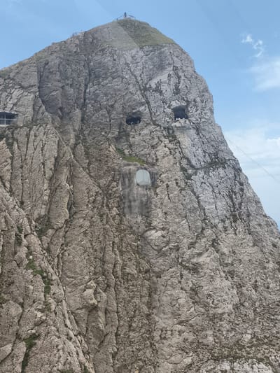 The image shows a sheer, rocky mountain face with two dark cave-like openings near the top and a circular window embedded in the rock face lower down. There are some people visible on the summit of the mountain, and a railing structure is present on the left side of the mountain. A cable wire stretches diagonally across the upper right portion of the image. The environment is mountainous and appears to be a tourist attraction given the presence of structures and people. The image depicts Mount Pilatus, accessible from Alpnach, Switzerland via a cogwheel railway, which is the steepest in the world, and a cable car.