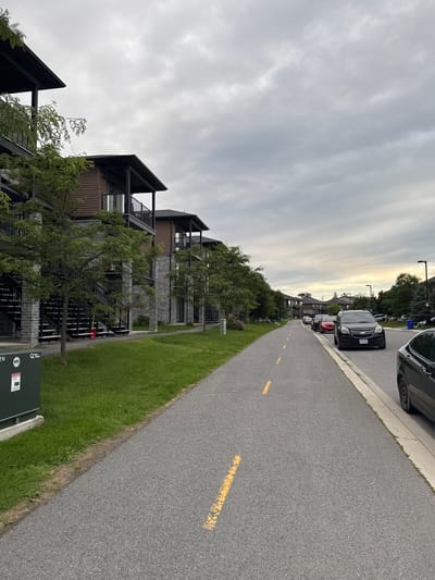 The image depicts a cloudy day scene along a paved pathway marked with dashed yellow lines, likely a bike or pedestrian lane, running alongside an apartment complex. The complex features multi-story units with brown siding and brick accents, external staircases, and balconies. Trees and grass are present between the pathway and the buildings. Several cars are parked on the right side of the pathway along a curb, with a black SUV prominently visible in the foreground. The environment appears to be a residential or mixed-use urban area in Gatineau, Canada. There are no people visible in the image.