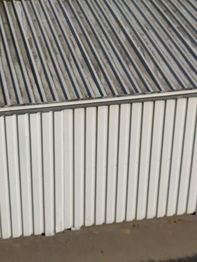 The image shows a close-up, aerial view of a corrugated metal roof and the upper section of a white corrugated metal wall. The roof, which occupies the upper portion of the image, is a light gray with dark gray vertical stripes and appears weathered. The wall, which is in the lower portion of the image, is white with thin, dark gray vertical seams and has a metallic sheen. The wall appears to be attached to a building. The ground below the wall is a light brown, textured surface, possibly dirt or asphalt. There are no people or animals visible in the image. This scene is likely an outdoor structure.