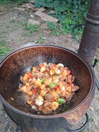 In Райымбек, Kazakhstan, a large, weathered cast-iron kazan is filled with a hearty stew of beef, potatoes, onions, and bell peppers. The food is visibly cooking, with a glossy sauce coating the ingredients. The kazan rests on a metal stand outdoors, with dry grass and dirt visible on the ground. In the background, lush green foliage and some scattered broken tiles can be seen. A tall, rusty metal pipe, possibly part of a chimney or stove, stands to the right of the kazan. The scene suggests an outdoor cooking or preparation setting, likely a meal being made for a group.