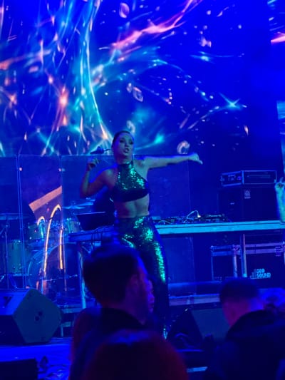 A female performer in a sparkling green outfit is on stage, dancing in front of a large, illuminated backdrop. She has her arms outstretched, and her mouth is open as if singing. Behind her, musical equipment, including a DJ mixer and speakers, are visible. The stage is bathed in blue light, with streaks of pink and white light emanating from the backdrop. In the foreground, out-of-focus audience members are visible, also illuminated by the stage lights. The event appears to be a concert or performance in Akhalubani, Georgia.