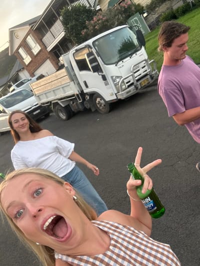 The image displays three individuals on an asphalt residential street in Thirroul, Australia, during what appears to be late daylight or early evening.

In the foreground, a blonde female, wearing a brown and white checkered top, is captured in a selfie perspective with an open-mouthed, wide-eyed expression directed at the camera. Her right hand, adorned with rings, grips a green glass bottle labeled "HAHN SuperDry," with her index and middle fingers extended in a "V" shape above the bottle's neck.

Slightly behind and to her left, a second female with long brown hair, wearing a white off-the-shoulder top and blue jeans, stands with a subtle smile directed towards the camera.

Further in the background and to the right, a male with medium-length brown hair, clad in a light purple t-shirt, is visible in profile, looking to his right, away from the camera.

Along the street, a white Isuzu NLR truck with a wooden-sided flatbed body is present. Its Australian license plate, "BC 08 SH," is partially discernible on the front bumper, which also features a silver bull bar. A white passenger vehicle is partially visible behind the truck. Residential brick buildings featuring white upper stories and balconies line the background, interspersed with various green vegetation and flowering shrubs. The sky exhibits a light orange tint, consistent with a dusk or dawn setting.