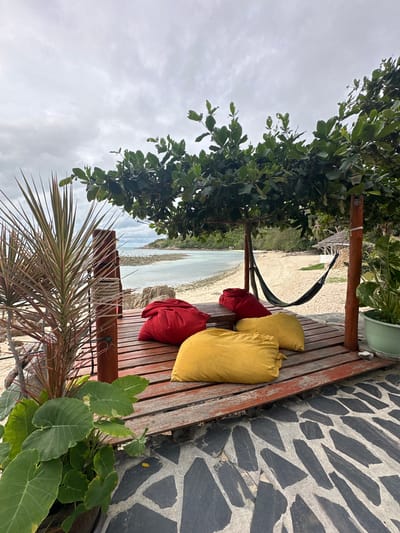 This is a photograph taken outdoors in the Ko Pha-ngan District, Thailand. The scene is a wooden deck situated near a beach. On the deck are several large, colorful floor cushions, two red and two yellow. A hammock is strung up between two wooden posts, partially shaded by the leaves of a large tree. Beyond the deck, a sandy beach meets a body of calm, light-colored water. The sky is overcast with gray clouds. In the foreground, a potted plant with large green leaves and a spiky plant with reddish-green leaves are visible. The ground in the foreground is paved with irregular, dark gray and light gray stones.