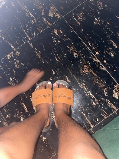 A pair of feet wearing tan sandals with white painted toenails are centered in the image. The right foot is in focus, while the left foot appears blurry suggesting movement. The footwear is on a dark, patterned tile floor. The sandal band has a darker signature in cursive across it. An additional leg extends into the left side of the image. A green surface is visible at the upper right corner of the scene.