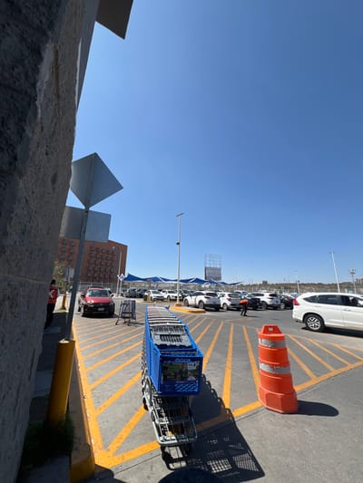 This image depicts an outdoor scene at a commercial parking lot in León, Mexico, under a clear blue sky. In the immediate foreground, a stack of approximately 10-12 blue shopping carts is visible on a cart return bay, positioned on concrete paving marked with yellow diagonal lines. To the right of the carts is a large orange traffic cone with two white reflective bands. On the far left, a textured grey concrete wall or pillar partially frames the shot, with a yellow bollard and a portion of a person wearing an orange vest visible near its base.
In the mid-ground, a red SUV is parked near another cart return bay. Further into the parking lot, numerous vehicles, predominantly white and light-colored SUVs and sedans, are parked beneath blue fabric shade structures. One individual wearing a dark uniform is visible near these parked vehicles, appearing to bend over or interact with the ground or a vehicle. In the far distance, a tall, multi-story building with reddish-brown facade and numerous windows is visible on the left. Several streetlights punctuate the scene, and a large metal billboard structure is discernible in the background on the right, beyond the shaded parking area. The ground consists of asphalt with yellow painted lines for parking demarcation.