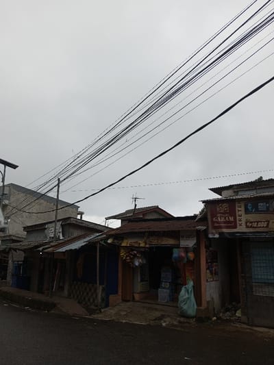 The image presents a street scene in Pacet, Indonesia, captured under a uniformly overcast sky. The foreground features a dark asphalt road, bordered on the left by a low, weathered concrete curb. Extending along the right side of the road is a row of contiguous, one-story commercial structures or residences, characterized by corrugated metal roofs, some exhibiting rust. Multiple black electrical and telecommunication wires are densely strung across the upper portion of the frame, originating from out-of-frame poles and a visible utility pole on the far left, which is equipped with a solar panel array.
No individuals are visible within the frame. The leftmost structure appears to be a multi-story building with unpainted concrete walls. The commercial establishments along the right have open shopfronts. One prominent shop on the far right displays a large banner advertising "Gudang Garam International KRETEK" with a price of "Rp 18,000" and an image of a man. Adjacent to it, another shop exhibits various packaged goods, including snacks and other items, hanging near its entrance, with a light green plastic bag resting on the ground directly in front. Further left, additional small structures with similar corrugated roofs are visible. Several rooftops feature small television or radio antennas. Minor debris and refuse are scattered near the base of the rightmost shop. No specific actions or interactions are discernible in the static scene.