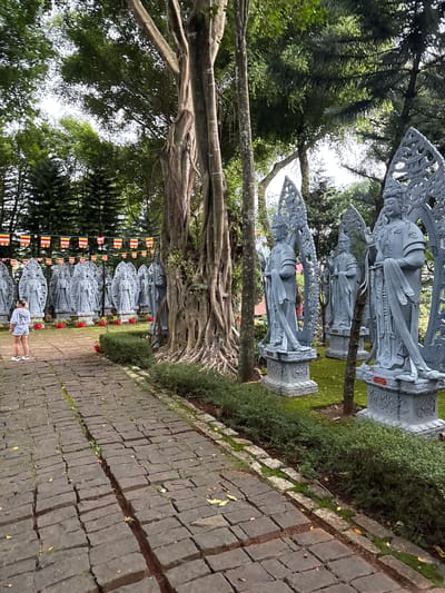 In Nam Ban, Vietnam, a woman stands on a brick pathway in a park filled with numerous grey statues of figures. The statues are lined up in rows, each on a pedestal and adorned with ornate headwear or halos. In the background, several strings of colorful Buddhist prayer flags are strung between trees. A large tree with a thick, gnarled trunk is prominent in the foreground. The ground is covered in moss and scattered yellow leaves. The overall scene depicts a serene, spiritual outdoor setting within a park.