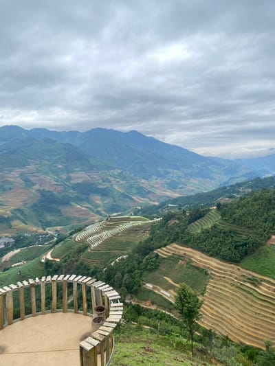 The image depicts a panoramic view of terraced rice fields on a mountainside, characteristic of the Mu Cang Chai region in Púng Luông, Vietnam. In the foreground, a circular observation deck with a rustic wooden railing provides a vantage point. The deck is made of concrete with several wooden posts and planks forming a fence-like structure. Two cylindrical planters, appearing to be made of woven material, are positioned on the deck. Beyond the deck, the landscape unfolds into a series of meticulously carved rice terraces that follow the contours of the rolling hills. The terraces display varying shades of green and brown, with some filled with water, reflecting the overcast sky. Lush green vegetation, including trees and shrubs, dots the slopes between the terraces. In the distance, more layers of mountains, shrouded in a soft haze, stretch towards the horizon under a cloudy sky. The scene conveys a sense of agricultural beauty and the harmonious integration of human activity with the natural environment.