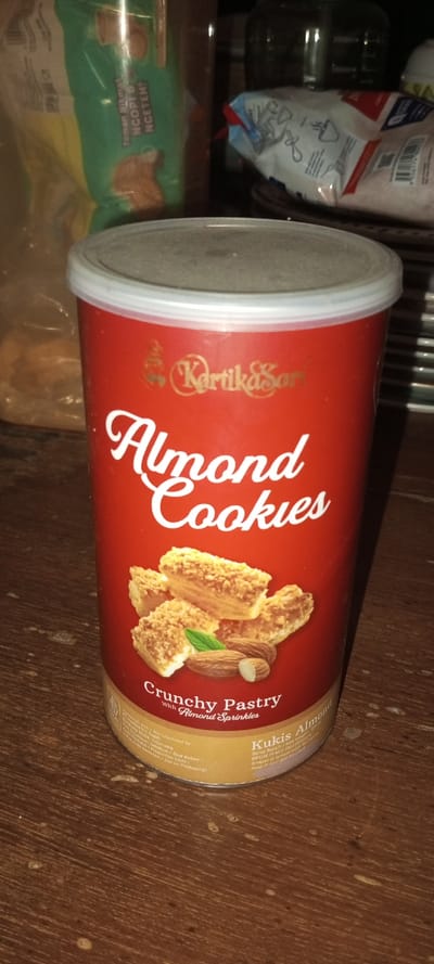 The image captures a red cylindrical container of "KartikaSari Almond Cookies" standing upright on a wooden surface, likely indoors. The container features the brand name "KartikaSari" in golden script, followed by "Almond Cookies" in prominent white cursive lettering. An illustration on the label shows several pieces of flaky pastry with sprinkles, alongside whole and shelled almonds. Below this, the text reads "Crunchy Pastry with Almond Sprinkles." Further down, partially visible text includes "Kukis Almond," "Diproduksi oleh / Manufactured by:", and "Indonesia," indicating the product's origin. Other fragmented text mentions "Berat Bersih / Net Wt:", "BPOM RI MD," and storage instructions "Simpan di tempat kering dan sejuk / Keep in cool and dry place."
In the blurred background, a green and yellow package is visible to the left, displaying Indonesian text "Teman Akrab NGOPI & NGETEH!" (meaning "Close Friend for Coffee & Tea!"). To the right, a white package and a clear glass jar are also present. The wooden surface shows some minor stains or watermarks.
The scene depicts a static arrangement of food items, possibly in a kitchen or pantry setting. There are no people or specific activities shown. The indoor lighting does not suggest a particular time of day. While the provided location context is Aral, Kyrgyzstan, the visible text on the packaging strongly suggests an Indonesian origin for the products, offering no direct visual cues aligning with Kyrgyzstan.