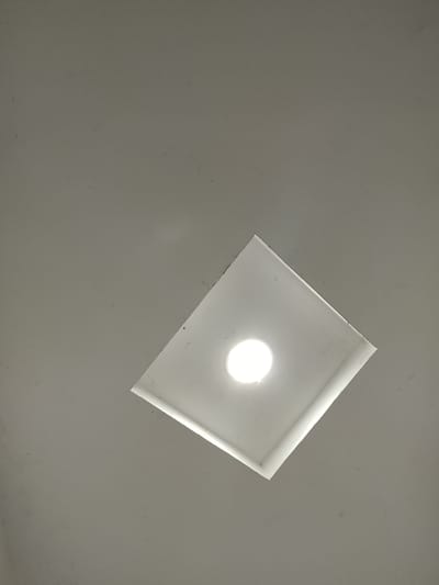 The image shows a close-up, high-angle shot of a ceiling in Rumuepirikom, Nigeria. A square-shaped recessed light fixture is visible, with a bright, circular light source at its center. The ceiling itself is a plain, neutral color, possibly white or light gray, and appears slightly textured. There are a few faint dark specks scattered across the surface, likely dust or minor imperfections. The lighting suggests the room is indoors, and the angle indicates the photograph was taken looking directly upwards. There are no people or other objects in the frame besides the ceiling and the light fixture.