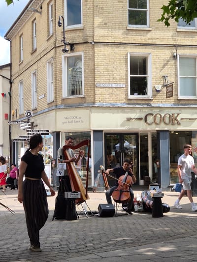 This is a medium shot, eye-level, outdoor photograph of a street scene in Bury St Edmunds, West Suffolk, United Kingdom. The main subjects are a cellist and a harpist performing on a cobblestone street in front of a building with a COOK store.

In the foreground, a young woman is playing the cello, seated on a chair. She is wearing a dark t-shirt and dark, striped, loose-fitting pants. Her instrument is positioned between her legs, and she is holding the bow to the strings. To her left is a black speaker. Next to her right is a laptop and a small donation box labeled "Thank you for your support." Behind the cello and donation box are various bags and equipment.

Slightly behind and to the right of the cellist, an older man is playing a harp. He is wearing a light grey t-shirt. He is seated on a stool and is actively plucking the harp strings with his hands. The harp is a large, wooden instrument.

To the left of the performers, a woman with dark hair is walking away from the camera. She is wearing a black crop top and striped pants similar to the cellist's. She is looking back over her shoulder towards the performers. Further to the left and in the background, other individuals are visible, including a woman in a white top and red skirt, and a woman in a pink top.

The building behind the performers has a light yellow brick facade and features several windows. A sign above the COOK store reads "We COOK so you don't have to." The COOK store entrance consists of glass doors. A street sign is visible on the building, indicating "BRENTGOVEL STREET."

The lighting suggests it is daytime, with sunlight casting shadows on the cobblestone street. The environment appears to be a pedestrianized area of a town or city.