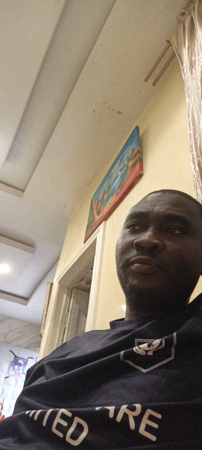 A low-angle shot depicts a dark-skinned man in a navy blue shirt. "UNITED WE" is visible in white lettering on the lower part of the shirt. His gaze is directed slightly upward and to the right. Behind him, a beige wall hosts a painting in a colorful frame and a doorway. A partial view of a patterned ceiling and white curtains adds depth to the scene. Overall, the lighting is soft, contributing to a relaxed and somewhat introspective atmosphere.