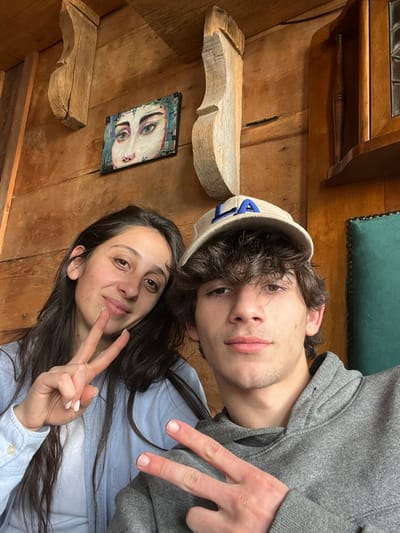 This indoor photograph captures two individuals, a female and a male, posing for a close-up image within a traditional interior setting in Argveta, Georgia.
The female subject is positioned on the left side of the frame, smiling faintly and looking directly at the camera. She has long, dark brown hair and is wearing a light blue button-up shirt over a lighter garment. Her left hand is raised, displaying a peace sign ("V" gesture).
The male subject occupies the right side of the frame, looking directly at the camera with a neutral expression. He has dark, curly hair partially covered by a light beige baseball cap with the letters "LA" embroidered in blue on the front. He is wearing a grey hooded sweatshirt. His right hand is raised, also displaying a peace sign ("V" gesture).
The background features wooden paneled walls. Mounted on the wall are two carved wooden corbels or brackets, with a small rectangular painting depicting human eyes and the upper part of a nose positioned between them. To the far right, a section of a dark green upholstered chair back with button tufting is visible, along with part of a wooden cabinet or shelving unit.