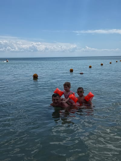 A man and two children are in the water at a beach in Yevpatoriya, Ukraine. The children are wearing orange inflatable armbands. A line of floating buoys extends into the water behind them. A few other people are visible in the distance, closer to the shore. The sky is blue with some clouds.