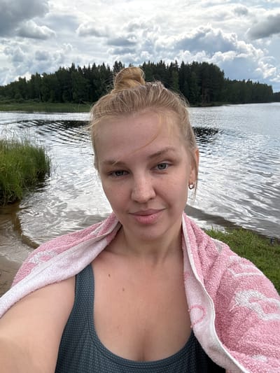 A young Caucasian woman with blonde hair pulled into a bun poses for a selfie outdoors. She is wearing a dark teal textured swimsuit and has a pink and white striped towel draped around her shoulders. She is standing near the water's edge, with a lake and a dense forest in the background under a cloudy sky. The location is identified as Zaymishche, Russia.