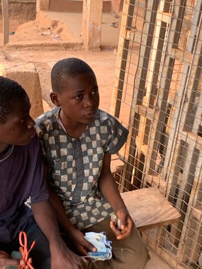 This image depicts two young males in an outdoor setting in Ube, Nigeria. The primary subject, a male estimated to be 8-12 years old, possesses short, dark hair and wears a short-sleeved collared shirt with a checkerboard pattern of light and dark grey squares. He is seated on a light-colored wooden surface. In his left hand, he holds a stack of various multi-colored banknotes. His gaze is directed towards the right side of the frame.
Positioned to his left and partially visible is a second male, also appearing to be 8-12 years old, wearing a dark blue short-sleeved top. This individual exhibits distinct linear scarification marks on his left cheek. He holds an orange braided cable in his left hand and a dark rectangular device in his right hand.
The background consists of exposed reddish-brown sandy earth and several weathered building structures, suggestive of aged concrete or mud-brick construction. To the right of the seated individuals, a tall, rectangular metal grid structure, possibly a gate or security screen, stands vertically, casting sharp linear shadows across the scene. The entire scene is illuminated by strong natural sunlight, creating a warm hue.
