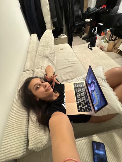 The image depicts a young woman lying on a beige sofa, using a laptop. She is wearing a black t-shirt and appears to be indoors, possibly in an apartment or dorm room. The background shows a cluttered space with clothing racks, furniture, and bags, suggesting a living area. There is also a smartphone on the sofa next to her. The overall lighting is dim. The location within the city of Mönchengladbach, Germany is not discernible from the image.