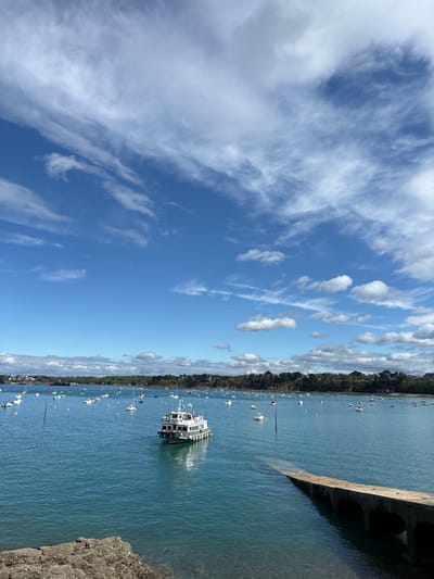 The image shows a wide-angle view of a harbor in Dinard, France, taken from a rocky outcropping at the water's edge. The sky is clear blue and dotted with wispy, white clouds. The water is calm and a bright turquoise color. There are several small boats and a large, white passenger boat in the water. The shore is visible in the background and is lined with trees. A concrete ramp leads down into the water from the rocky outcropping. There are no people visible in the image.