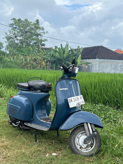 The image shows a blue Vespa scooter parked on a grassy field in Celuk, Indonesia. The scooter has a license plate that reads "DK 2426 KF". The background includes lush green rice paddies, a house with a brown roof, and banana trees. The sky is cloudy.