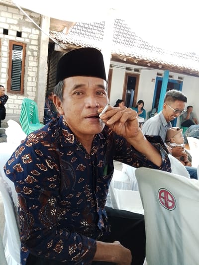 The image portrays an outdoor social gathering in Juwana, Indonesia, under bright daylight conditions. In the foreground, an adult male, appearing middle-aged or elderly, is seated. He wears a black peci and a long-sleeved dark blue batik shirt with brown and white patterns. He holds a lit cigarette to his mouth with his right hand, looking directly towards the viewer. A green pen is clipped into his left shirt pocket. He is seated on a white chair. To the immediate right, a portion of another white chair with a white fabric cover displays a circular red and white logo with an 'AB' monogram.
In the mid-ground, a man with short hair and glasses is seen smiling while holding an infant wearing a light brown cap. Further back, multiple individuals, including other men and women (one in a blue top), are visible, seated on white chairs. A light green plastic chair is also present on the left side of the mid-ground. The background features a building with a terracotta tiled roof, white walls, and windows, some with blue frames. A partially constructed brick wall with an exposed window is visible on the far left. An overhead white canopy structure provides shade to the gathering area.
