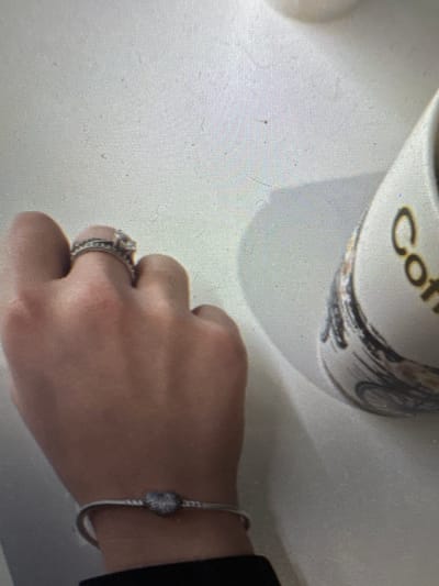 The image shows a close-up view of a person's left hand and wrist, adorned with rings and a bracelet. On the ring finger, two silver bands are visible, one with small stones and the other appearing to be a wedding band or engagement ring. A delicate silver bracelet with a heart-shaped charm, possibly encrusted with small blue or grey stones, is worn on the wrist. To the right of the hand, a portion of a white mug with the word "Coffee" visible in bold black lettering is present. The mug has a black, abstract design. The background is a plain, textured surface, likely a table or countertop. The image does not provide any specific details or context related to the city of Murcia, Spain.