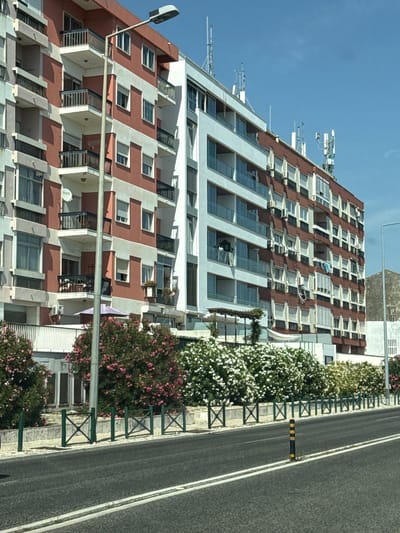 The image shows a street scene in Oeiras, Portugal, with a focus on a multi-story residential building. The building has a distinctive red and white facade, with numerous balconies and windows. Several air conditioning units are visible on the exterior walls. On the rooftop, a number of telecommunication antennas are installed. In front of the building, there is a line of dense flowering bushes, some with pink blossoms and others with white blossoms. A low green fence with decorative diamond patterns separates the sidewalk from the street. A lamppost stands prominently in the foreground. The street itself is paved, with double white lines marking the lanes. A yellow and black striped bollard is positioned on the side of the road. The sky above is clear and blue. The overall impression is a well-maintained urban environment on a bright, sunny day.