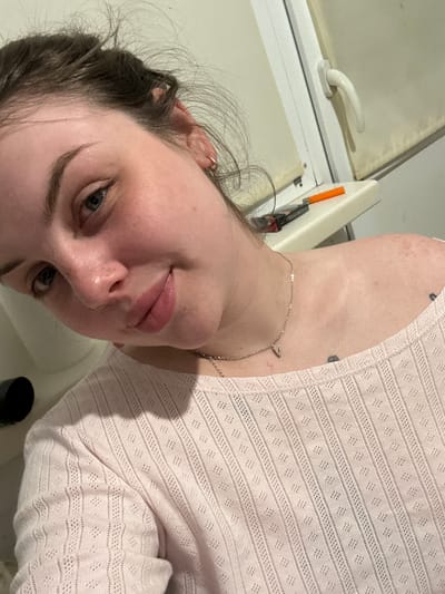 A selfie taken indoors features a fair-skinned person with dark hair pulled back, wearing a light pink open-weave shirt and a delicate gold necklace with a "V" pendant. The person is angled towards the camera with their head tilted. The background includes a white window frame with a handle, suggesting an interior space. The location in Chornomorskoe, Ukraine is not evident from the image.