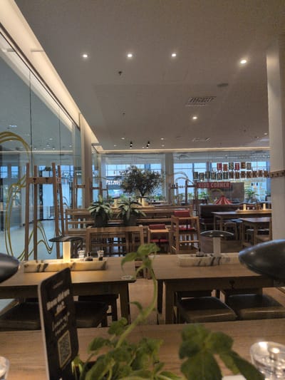The image shows an indoor dining area with wooden tables and chairs, likely at a restaurant. Several tables are visible, some set with glasses and condiments. In the background, through a large glass partition, a shopping mall interior is visible with shops and escalators. There are also decorative elements like plants and a sign that reads "KIDS CORNER". The ceiling features recessed lighting and air vents. There are no people clearly visible in the scene. The overall impression is one of a modern, casual dining space within a larger commercial complex. The sign "LASTEN LEIKKINURKKAU" along with "KIDS CORNER" suggests this location is in Helsinki, Finland, likely within a shopping center such as Jumbo or Sello, which are known for having family-friendly amenities.