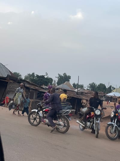 The image shows a street scene in Bokana, Nigeria. Multiple motorcycles are parked along a dirt road, with people seated on them or standing nearby. The background features simple, weathered buildings with corrugated metal roofs and trees. The sky is overcast. People are dressed in casual clothing. One man wears a yellow hat.