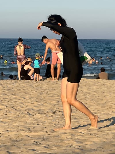 This media file depicts a beach scene in Đà Nẵng, Vietnam. The primary subjects are a group of people enjoying a day at the beach. In the foreground, a woman in a black full-body swimsuit is the most prominent figure, with her legs extended on the sand. Behind her, several other individuals are visible, some in swimwear. One man wears pink and blue striped swim shorts, and a child is seen in a blue and yellow shirt and black shorts, wearing a green cap. Further back, more people are in the water, and some are partially obscured by other beachgoers.

The setting is clearly a sandy beach with the ocean in the background. The water appears calm with gentle waves. The sand is textured, showing the typical patterns left by the receding tide or wind. The time of day appears to be late afternoon or early evening, as suggested by the soft, warm light casting shadows on the sand. The sky is clear and a pale blue, indicating fair weather.

The overall atmosphere seems relaxed and leisurely, with people engaging in typical beach activities such as sunbathing and wading in the water. There are no specific events clearly taking place, but rather individuals enjoying their time at the beach.

Notable details include the varying attire of the people, suggesting a casual and diverse group. The presence of what appears to be a beach umbrella or sail in the background, along with the clear sky and calm sea, reinforces the impression of a pleasant day at the coast. No visible text is present in the media file.