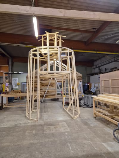 An interior scene captures a workshop environment in Leudelange, Luxembourg, where a partially constructed wooden structure dominates the center. The structure, resembling a tower or gazebo, is made of light-colored wood framing, with vertical posts and horizontal crossbeams forming its shape. Atop the main structure, a smaller, more intricate wooden frame is visible, suggesting a roof or an upper level.
The workshop floor is paved with square, grey tiles. In the background, the workshop walls are a muted green, with exposed wooden beams and metal framing supporting the ceiling. Fluorescent lighting fixtures are attached to the ceiling. Various tools and materials are scattered throughout the space. To the left, a workbench with wooden planks is present. In the middle ground, a silver ladder leans against a wooden wall. To the right, a large wooden workbench with wheels holds stacks of wooden planks and coiled cables. Other workshop equipment, including a large grey plastic container and what appears to be a piece of industrial machinery, are also visible. The overall impression is one of ongoing construction and a busy workspace.
