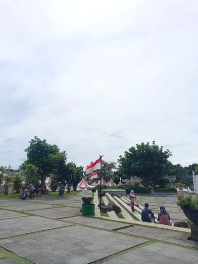 A wide, paved area with green grass growing in the cracks. Several people are sitting on stone steps, while others walk nearby. Indonesian flags and red and white triangular pennants are placed around the area. Trees are scattered throughout the scene under a cloudy sky. A couple are on bicycles on the left side. There are potted plants on both sides of the image.