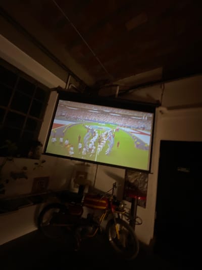 The image shows a dimly lit room where a projector screen displays a soccer match to an audience of people. A red and yellow moped is parked in the foreground. The room appears to be located in the city of Cucuron, France.