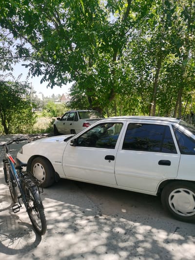 The image depicts a street scene in the city of Karabair, Uzbekistan. In the foreground, a blue and black bicycle is parked next to a white Daewoo Nexia sedan. The car is facing right, and its right side is visible. The bicycle is positioned on the left of the car, angled towards the viewer. The ground is paved with a rough, grey surface. Behind the white sedan, another white sedan, also a Daewoo Nexia, is parked. This second car is further back and partially obscured by trees. The background consists of dense green foliage and trees, suggesting a rural or suburban environment. The sky is visible through the leaves, indicating a clear day. The overall impression is a still, outdoor scene, likely on a residential street or a quiet public area.