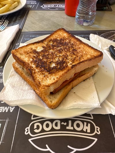A close-up, eye-level shot shows a toasted sandwich with a hot dog inside, served on a white plate with a white napkin. The sandwich is the main focus, with its golden-brown, crispy toast and visible filling. The plate rests on a black placemat featuring a white "HOT DOG" logo. In the background, a white napkin and a fork are visible on the left, while a red cup, a plastic water bottle, and a dark beverage are on the right. The setting appears to be an indoor eatery, likely a cafe or restaurant, in Sant Feliu De Guíxols, Spain, suggested by the Spanish text on the placemat.
