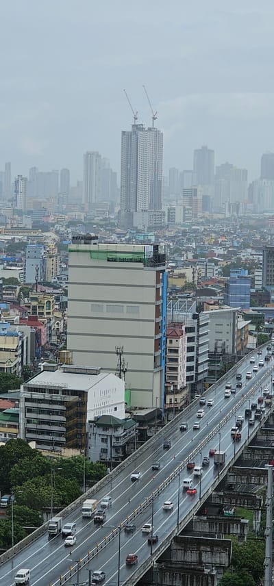 This is a high-angle, slightly hazy, vertical shot of an elevated highway in Makati City, Philippines, on a cloudy day. The highway, with multiple lanes, is wet, indicating recent rain, and is filled with various vehicles including cars, vans, and trucks. The road surface is reflective due to the moisture.

Dominating the mid-ground and left of center is a large, light-colored, multi-story building with horizontal gray stripes and a green facade on its upper section. To its right, a taller, slender building with a red roof and numerous windows stands out. The background is a dense urban landscape of many other buildings of varying heights and architectural styles, some appearing to be under construction with cranes visible. The overall impression is of a busy, sprawling metropolitan area.