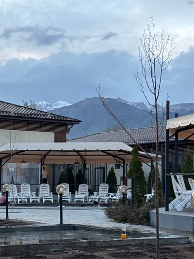 The image shows a street view in Kara-Oy, Kyrgyzstan. The scene is dominated by modern buildings with a mountain range visible in the background, partially obscured by clouds. In the foreground, there are rows of white plastic chairs along a paved walkway, interspersed with lampposts. The architecture includes brown and white facades with dark metal awnings. A bare tree branch extends into the upper left of the frame, contrasting with the sky. A few people appear to be relaxing on some of the chairs.