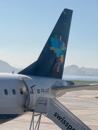An Embraer E-Jet series aircraft, registered PR-AXE, is depicted parked on an airport tarmac under clear, bright daylight conditions. The rear fuselage, tail fin, and a portion of the right wing are prominently featured. The aircraft's tail fin is dark blue and adorned with a colorful mosaic-style representation of the map of Brazil, composed of various rectangular shapes in shades of blue, green, yellow, red, and purple. The alphanumeric designator "XE" is visible near the top of the tail fin. The white fuselage displays the Brazilian flag emblem and the registration "PR-AXE" forward of the rear passenger door. This door is open, revealing a glimpse of the aircraft's interior and a partial "EXIT" sign. A mobile passenger staircase, branded "swissport" with its logo, is positioned against the open aircraft door, indicating active ground operations such as boarding or deplaning. The tarmac beneath is grey with yellow and red painted markings. In the background, a large body of water, likely Guanabara Bay, separates the airport from a series of hazy, tree-covered mountains and urban structures in the distance, characteristic of the city of Rio de Janeiro, Brazil.
