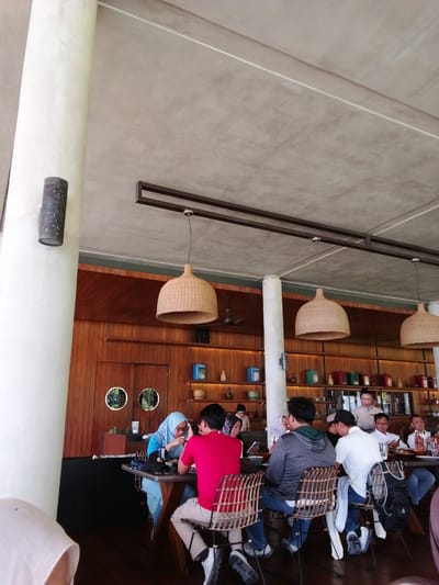 The image depicts the interior of a restaurant in Banyuwangi, Indonesia, characterized by a rustic and natural ambiance. A large, white pillar dominates the left side of the frame, reaching towards a textured concrete ceiling. Above, several large, woven pendant lamps hang, casting a warm glow. The background features a wall paneled with dark wood, adorned with shelves displaying various ceramic vases and decorative items.
Several patrons are seated at tables, engaged in conversations and meals. In the foreground, a group of men and women are seated around a table. One woman in a blue hijab is seated opposite a man in a red shirt. Further down the table, other individuals are visible, some interacting, others appearing to focus on their meals or phones. A server, a woman in a white uniform, is also seen interacting with guests at one of the tables. The overall atmosphere suggests a relaxed dining experience, possibly during daylight hours given the ambient light.