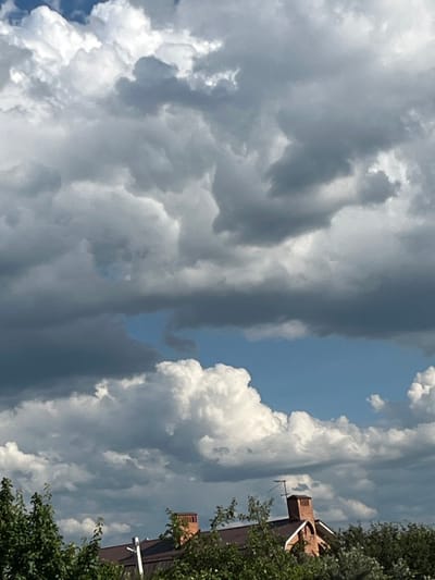 The media file is a photograph taken outdoors, likely during the daytime, given the bright sunlight illuminating the clouds. The scene is dominated by a dramatic sky filled with a mix of dark, heavy storm clouds and bright, puffy cumulus clouds, suggesting a dynamic weather system. A sliver of clear blue sky is visible between the cloud formations.
On the right side of the image, the upper portion of a building is visible, constructed of brick and featuring some architectural details like a pitched roof and a chimney. The building is partially obscured by lush green foliage from trees in the foreground. The presence of trees and a building suggests an urban or suburban setting.
The image is oriented vertically, making it appear as if the viewer is looking upwards at the sky and the building. There are no people or specific activities depicted. The weather appears to be unsettled, possibly transitioning between fair and stormy conditions. The location context of Prigorodny, Russia, places the scene in a specific geographical area. No visible text is present in the image.