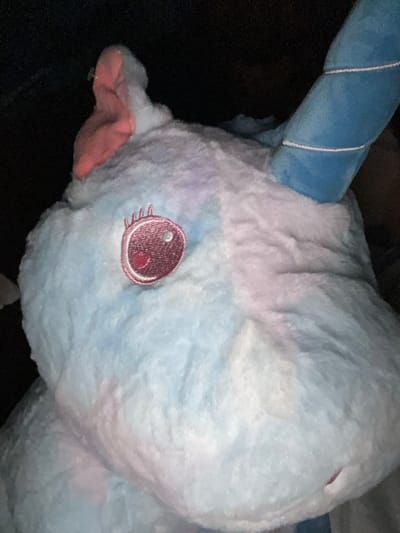 A close-up shot shows the head of a plush toy. The toy is primarily light blue and white, with a pink snout and a blue horn. The horn has white stripes spiraling around it. The toy has a large, embroidered eye with pink and red details. The background is dark and indistinct. The location is unknown and not discernible from the image. The city of Kaloyanovo, Bulgaria, cannot be connected to the image based on available visual information.