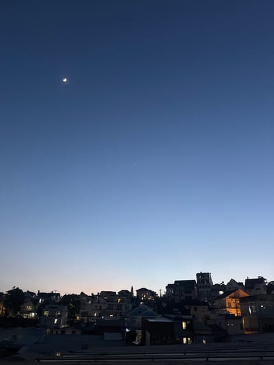 The image captures a twilight scene over Lam Vien - Da Lat, Vietnam, showcasing a dense urban landscape against a gradient sky. The sky transitions from a deep, dark blue at the top to a lighter, softer blue near the horizon. A sliver of the moon is visible in the upper left quadrant, and a single, distant star or planet is present slightly to the right of the center.
The foreground is dominated by rooftops of various buildings, some with visible corrugated metal and others appearing as darker, flat surfaces. Beyond these, a hillside is covered with a dense arrangement of houses and buildings. These structures display a mix of architectural styles, with pitched roofs, some with dormer windows, and others featuring flatter, more modern designs. Many of these buildings have illuminated windows, indicating indoor activity as dusk settles. The houses are built closely together, suggesting a hilly and perhaps crowded urban environment.
The overall impression is one of a city winding down for the evening, with the last light of day illuminating the sky and the moon and stars beginning to emerge. The perspective is looking upwards towards the hillside and sky from a slightly elevated position. No people are visible in the frame.