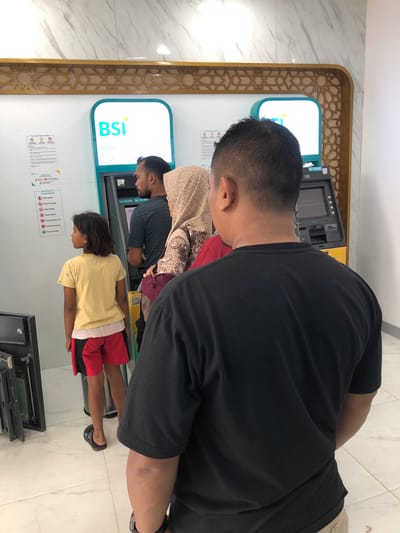 This is a photograph taken indoors, likely in a bank or ATM lobby, as indicated by the presence of an ATM machine and signage. The scene shows at least four people waiting in line to use the ATM. The first person in line is a young child wearing a yellow t-shirt and red shorts, facing the ATM. Behind the child is a man wearing a dark shirt and a head covering. Further back, another man in a black t-shirt is visible, with his back to the camera. The ATM machine itself displays the "BSI" logo, which stands for Bank Syariah Indonesia. Text on the ATM screen indicates "setor Link 1" and displays currency denominations of Rp 100,000 and Rp 50,000. Informational posters are visible on the wall near the ATM, including one titled "Do & Don'ts Uang Disetor" which lists instructions for depositing money. The overall setting appears to be a clean, well-lit interior with tiled flooring and marble-patterned walls. The time of day is not explicitly stated but the lighting suggests it is daytime. The individuals appear to be calmly waiting their turn. The image is oriented vertically.
