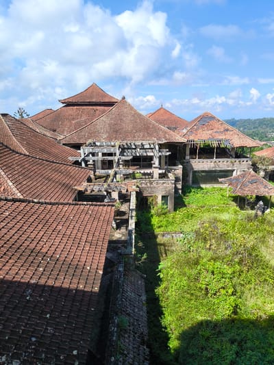 The image depicts an abandoned resort in Baturiti, Indonesia, under a bright blue sky with scattered white clouds. The resort features multiple buildings with traditional terracotta tiled roofs, many of which are in a state of disrepair with missing or broken tiles, revealing the wooden structures beneath. The architecture appears to be Balinese-inspired, with multi-tiered roofs and open-air structures.
The resort is overgrown with lush green vegetation, with trees, bushes, and vines encroaching on the buildings. The once manicured grounds are now wild and untamed. The buildings themselves show signs of decay, with crumbling concrete, broken windows, and peeling paint. Some areas of the resort seem to have been designed with courtyards or water features, now filled with foliage.
The overall impression is one of abandonment and nature reclaiming the man-made structures. The scene is still and silent, with no people visible. The surrounding landscape suggests a hilly or mountainous terrain, with distant hills covered in greenery. The image captures a sense of lost grandeur and the passage of time.