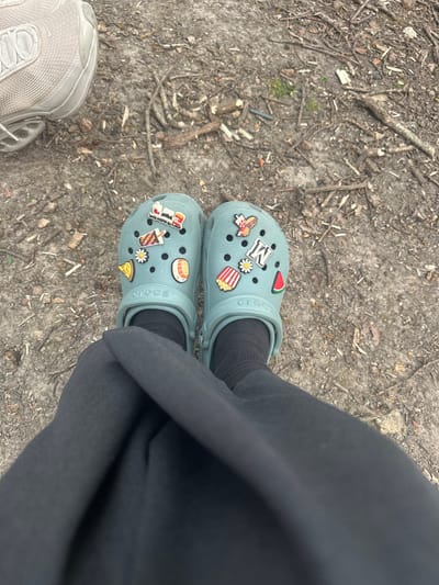 The image shows a ground-level view of a person wearing light blue Crocs with various charms in the holes. The person is also wearing black socks and black sweatpants. A single gray sneaker is visible at the bottom left of the frame. The ground appears to be dirt and twigs. Based on the location (Châtenay-Malabry, France), it is likely a park or wooded area in the city.