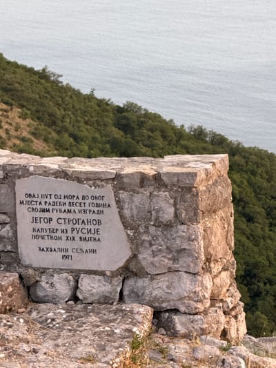 The image depicts a stone monument with an inscription, situated on a rocky outcrop overlooking a body of water and a densely forested hillside. The monument is constructed from rough-hewn stones, with a prominent plaque bearing text in Cyrillic script. The inscription reads, "This path from the sea to this place was built over ten years by his own hands by Jegor Stroganov, a monk from Russia in the early 19th century. Grateful villagers 1971." The plaque is partially damaged, with a crack running across its surface. The environment is natural and rugged, suggesting a remote or historical location. The sea in the background appears calm and vast, while the hillside is covered in lush green trees. The lighting suggests it is late afternoon or early evening, with a warm glow illuminating the stone monument. The monument is located in the village of Čelobrdo, Montenegro, a place known for its scenic views and historical significance.