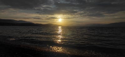 The image captures a serene sunset over the sea, with the sun peeking through a dramatic, cloudy sky. The sun's rays create a shimmering reflection on the dark, rippling water. In the foreground, a pebble beach is visible, with the gentle waves lapping at the shore. Silhouetted mountains form a distant horizon line, adding depth to the landscape. The overall mood is peaceful and tranquil, characteristic of the Municipal Unit of Aegio, Greece, a coastal region known for its beautiful sunsets.
